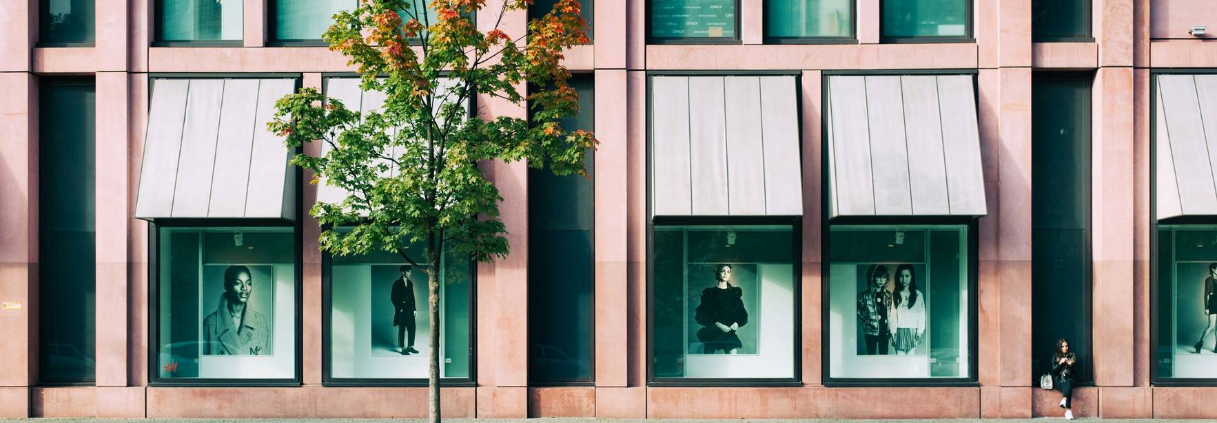 La imagen muestra una fachada de un edificio con fotografías de moda en sus ventanas y un árbol en la acera.
