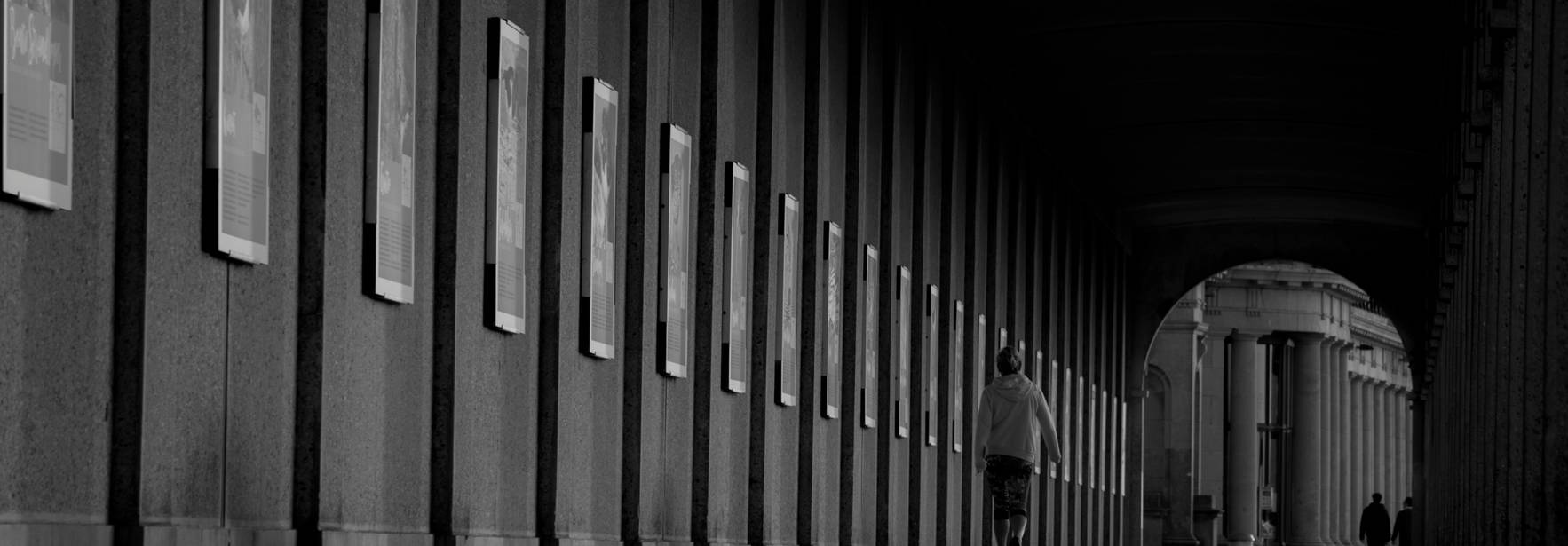 Un pasillo con columnas y carteles en las paredes, donde se observa a varias personas caminando en un ambiente en blanco y negro.