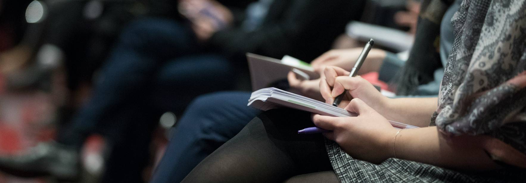 Un grupo de personas sentadas atendiendo a un evento, tomando notas en sus cuadernos.