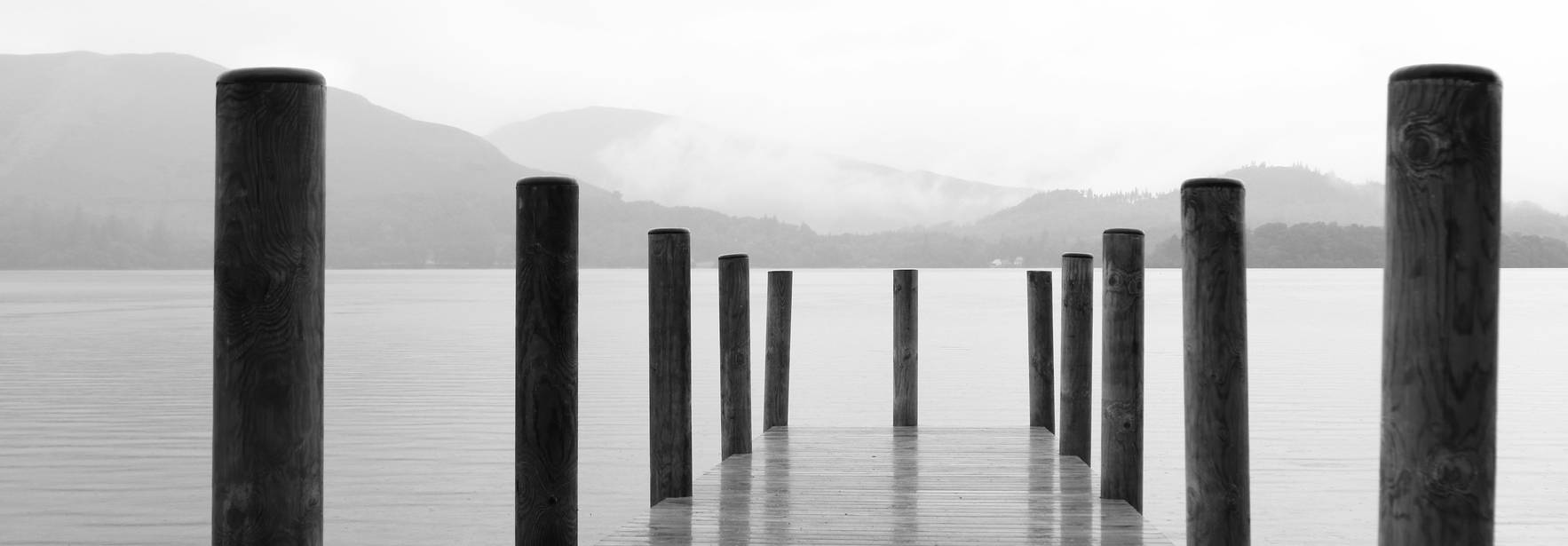 Una pasarela de madera se extiende sobre un lago tranquilo, rodeada de columnas y un ambiente sereno y nublado.