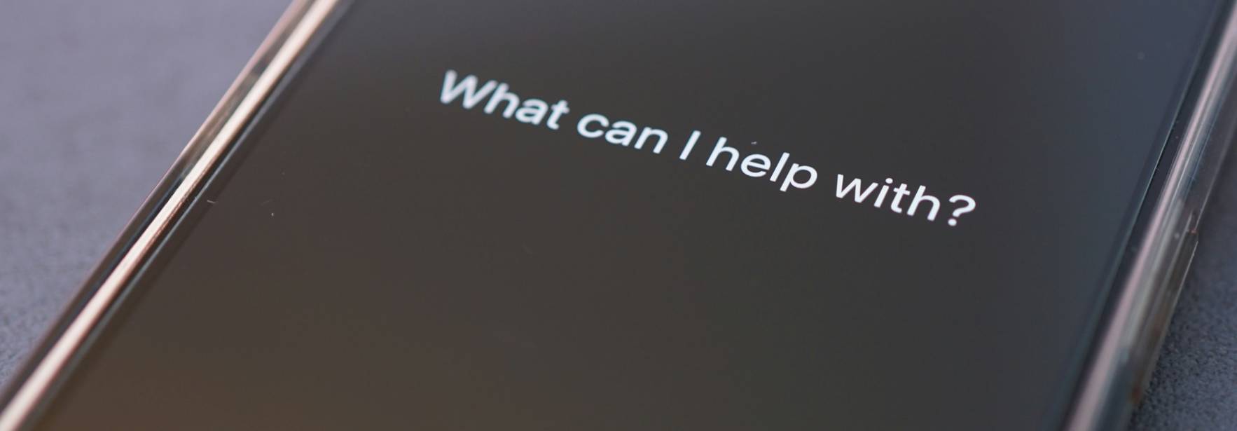La imagen muestra un teléfono móvil con una pantalla oscura que tiene un texto en inglés que pregunta cómo puede ayudar.