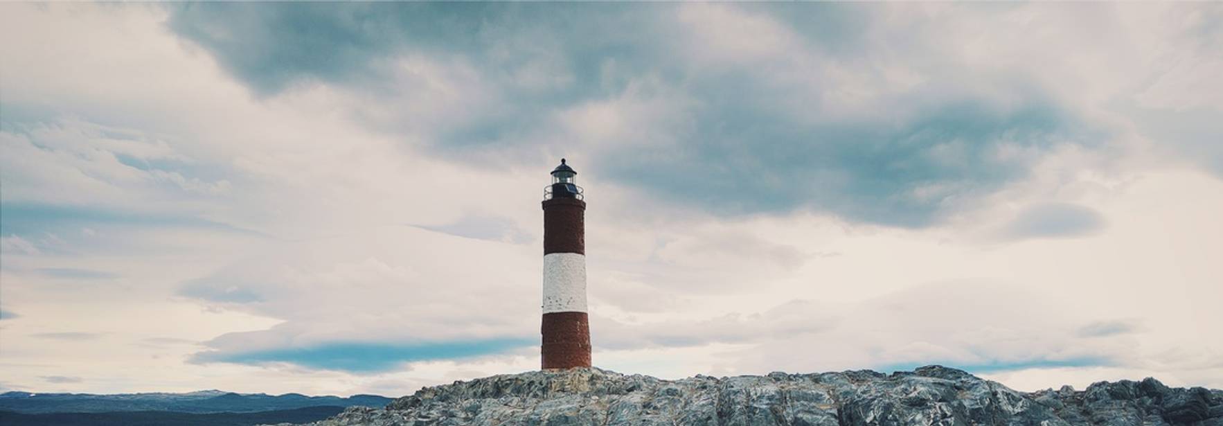 Un faro se erige sobre una formación rocosa en medio del océano bajo un cielo nublado.