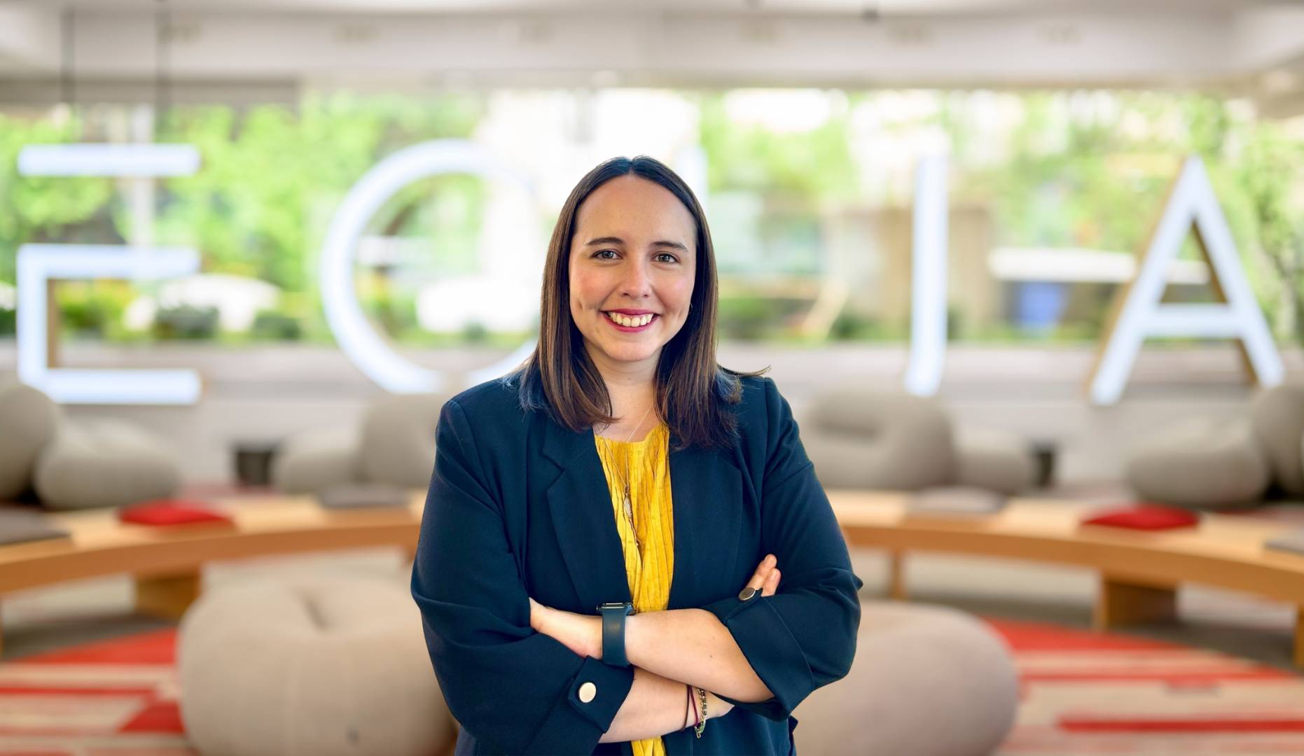 Mujer sonriente de pie frente a un fondo de letras iluminadas en un espacio moderno.