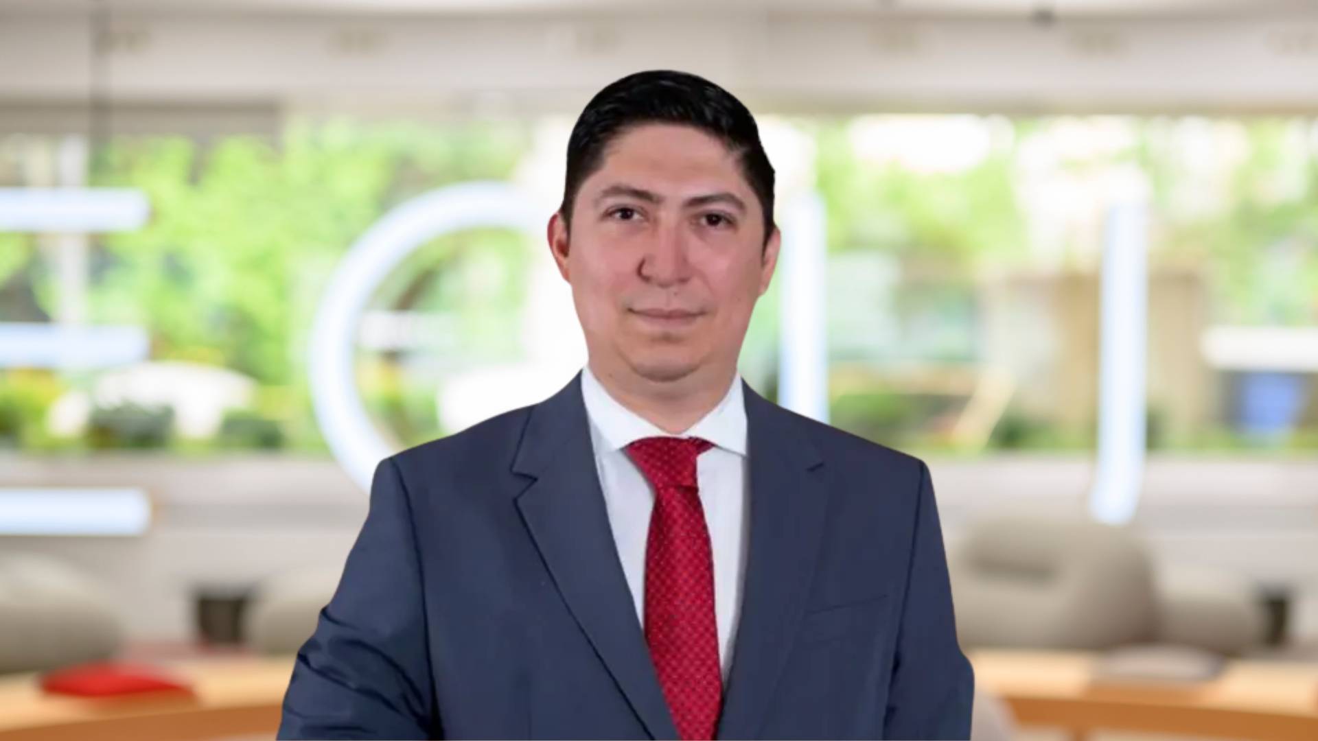 Un hombre vestido con traje y corbata sonríe frente a un fondo moderno de oficina.