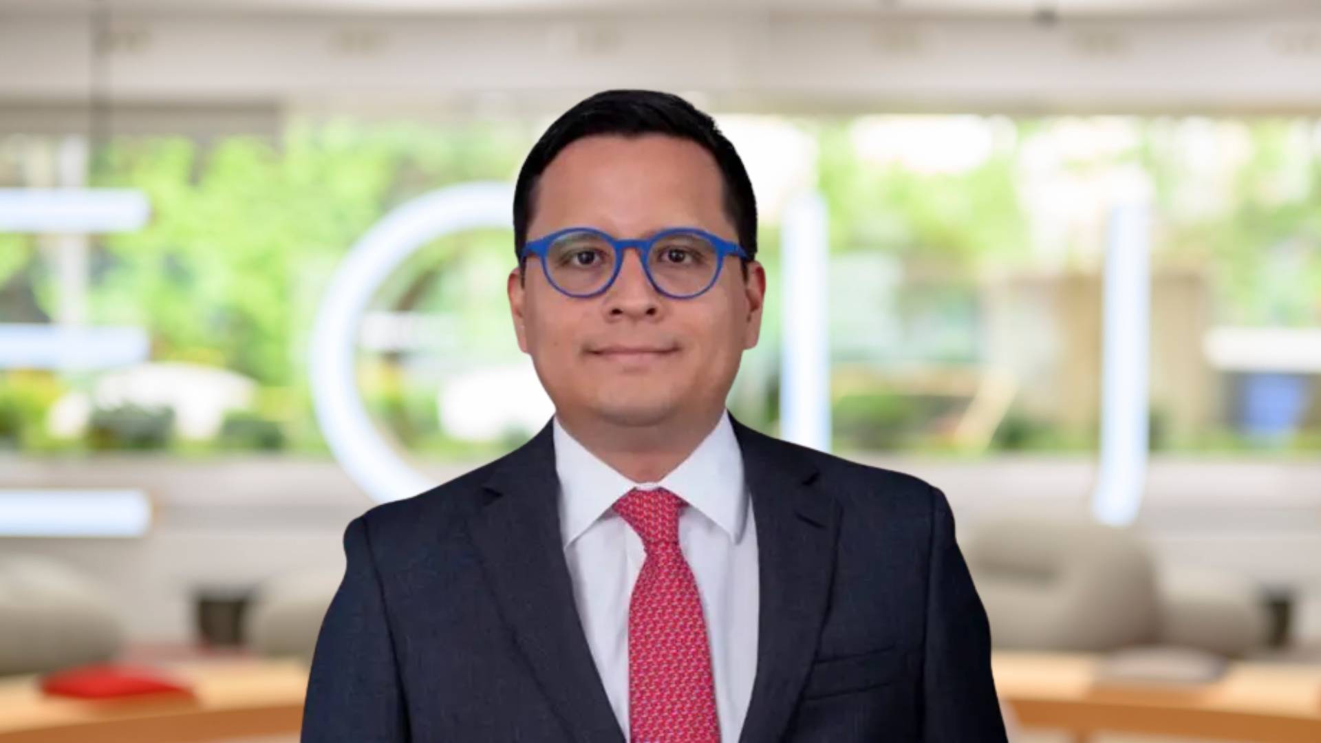 Una persona de pie frente a un fondo moderno y desenfocado, vistiendo un traje y lentes.