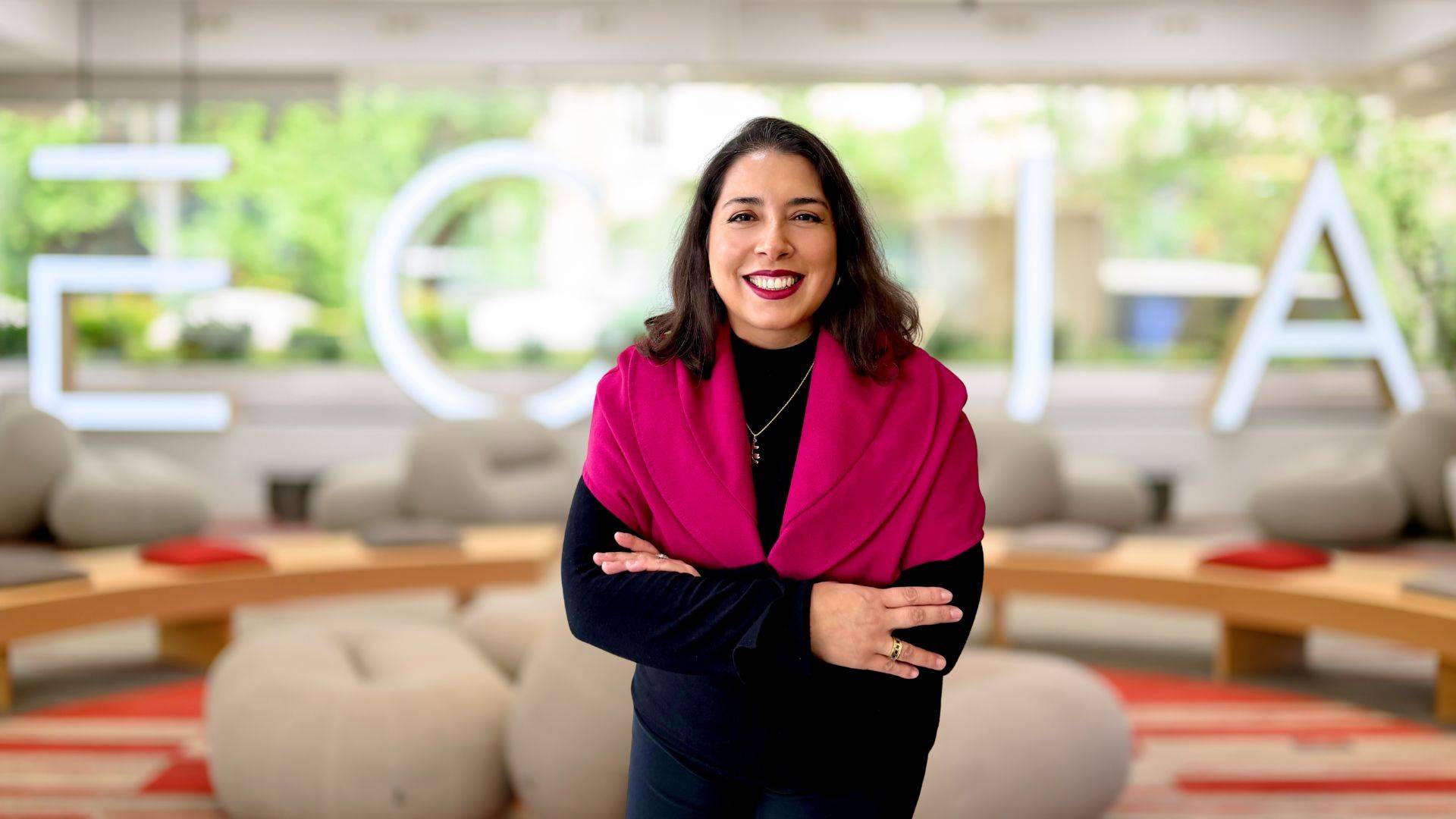 Una mujer sonriente se encuentra de pie con los brazos cruzados, vistiendo un chal fucsia, en un entorno moderno y acogedor.