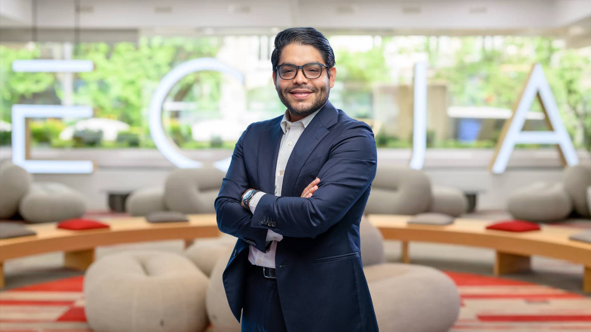 Un hombre sonriente con gafas y traje negro se encuentra de pie en un ambiente moderno y acogedor.