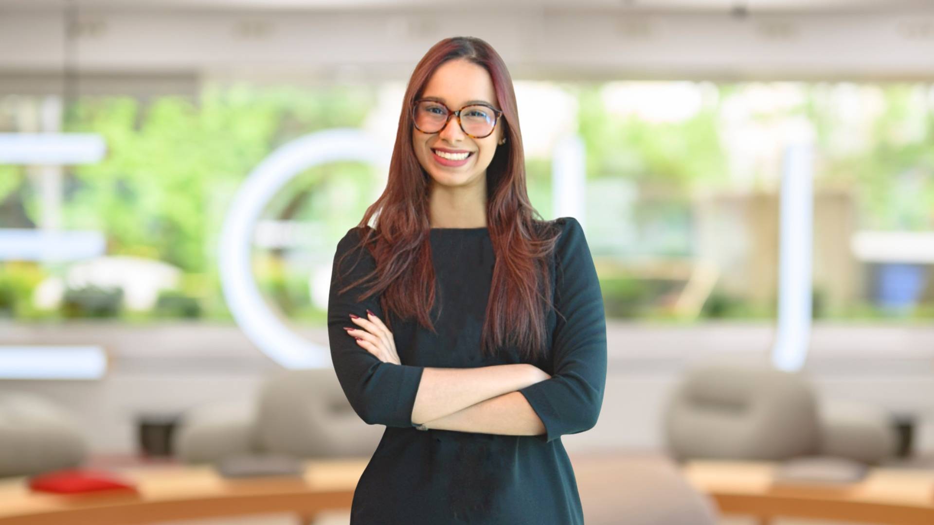 Una mujer sonriente con gafas de vista está de pie con los brazos cruzados en un entorno de oficina moderno.
