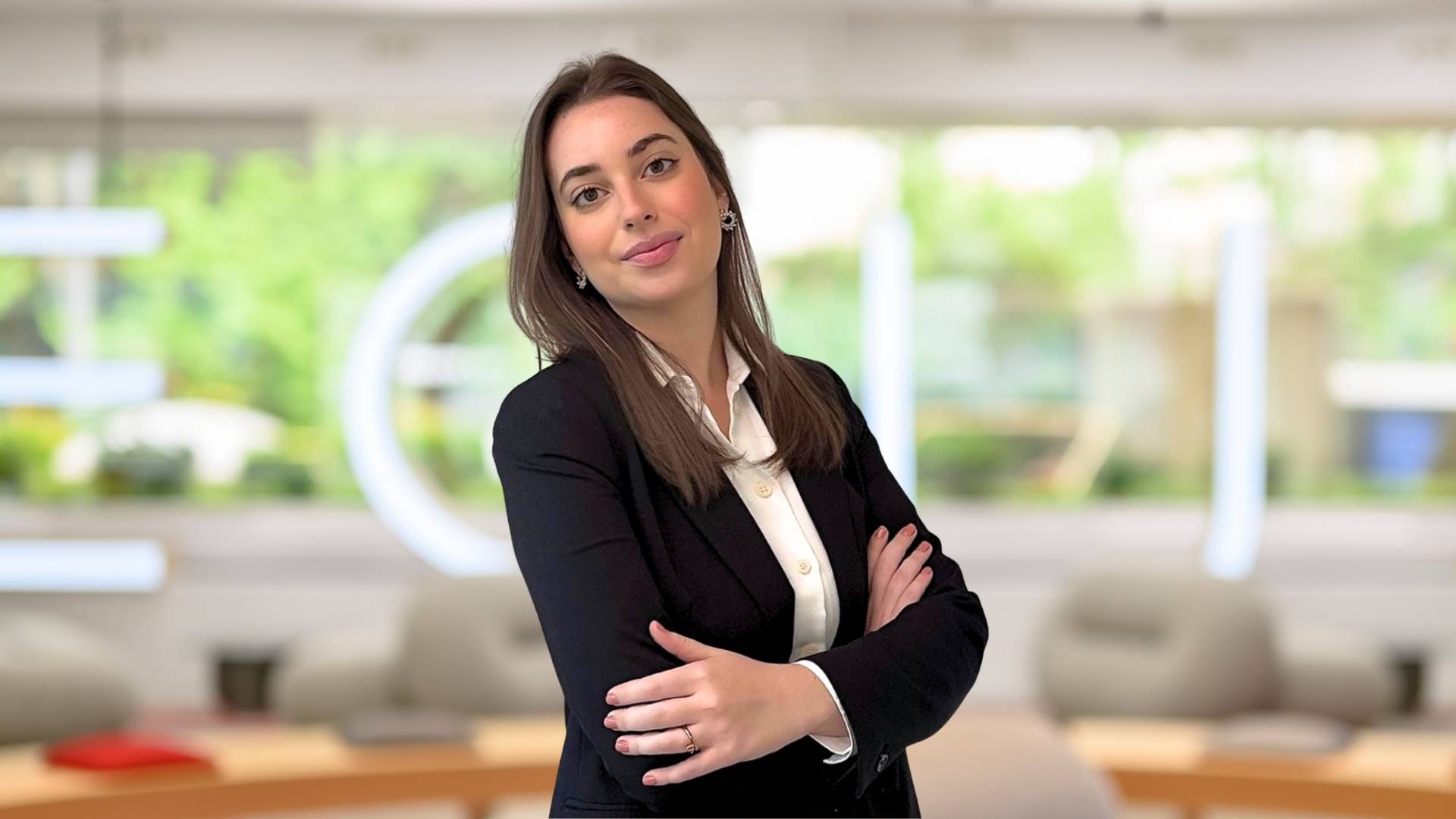 Una mujer sonriente viste un traje negro con una blusa blanca y se encuentra en un ambiente de oficina moderna.