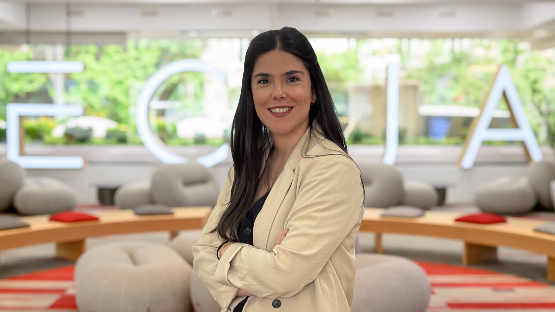 Una mujer sonriente con un blazer claro se encuentra en un entorno moderno y luminoso.