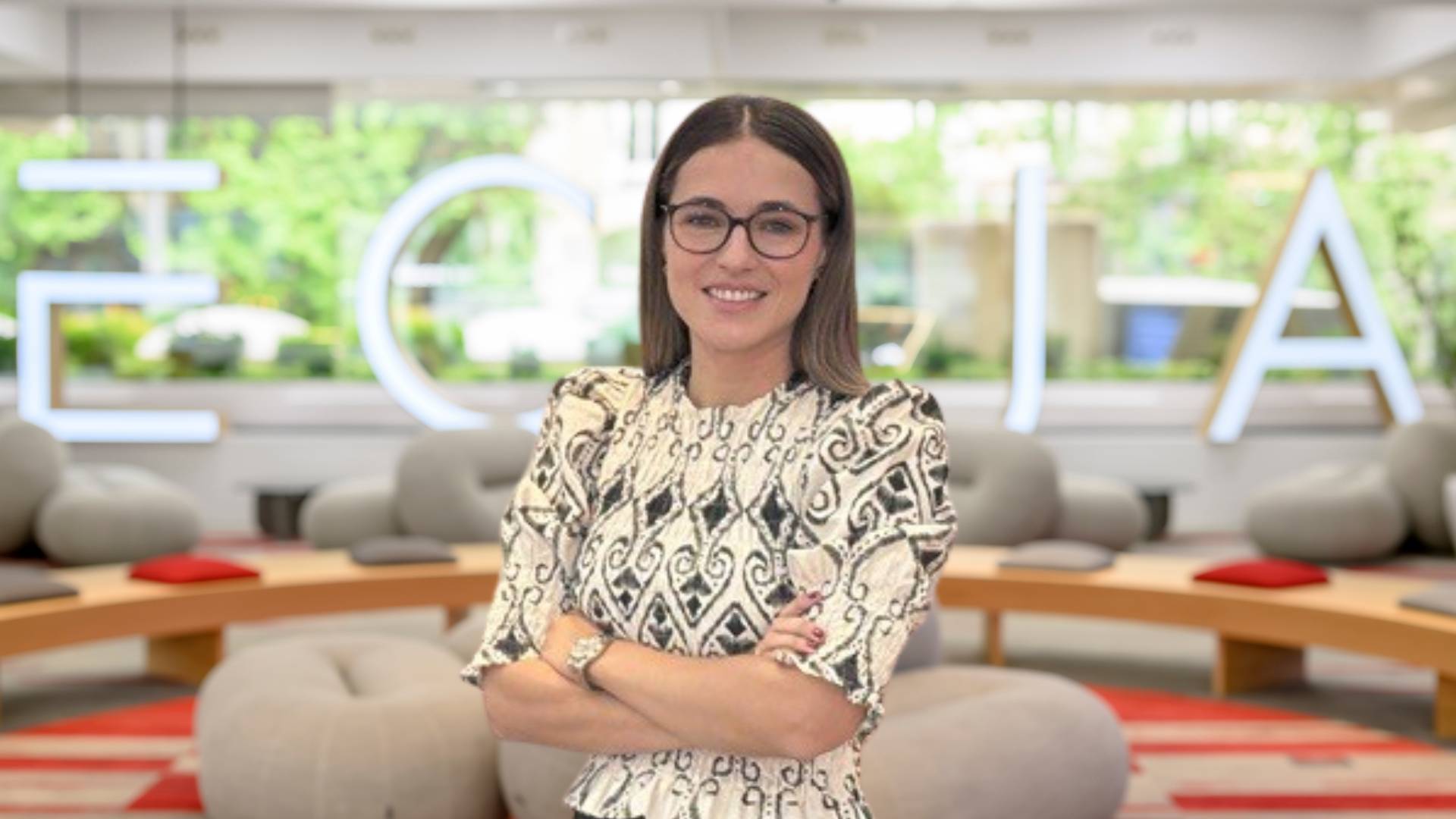 Una mujer sonriente con gafas y un vestido de diseño elegante posa frente a un fondo moderno con el nombre de una empresa iluminado.