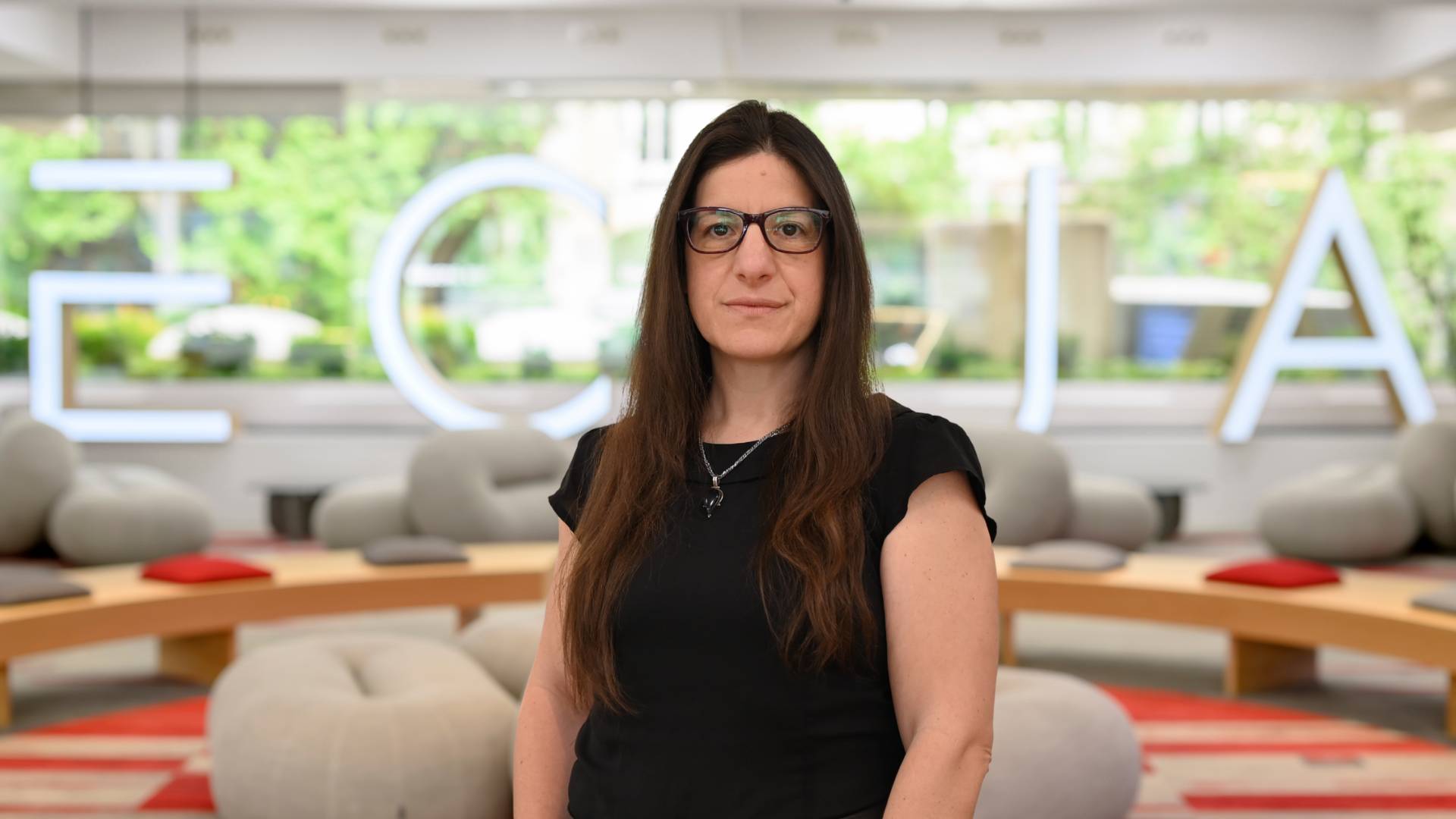 Una mujer de cabello largo y lacio, con gafas, posa frente a un ambiente moderno y luminoso con el fondo de una palabra en gran formato.