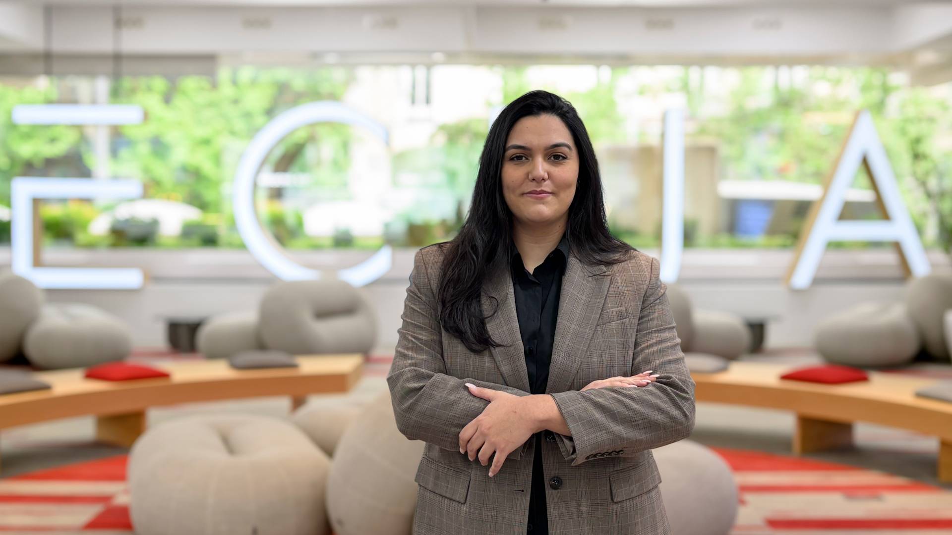 Una mujer de pie en un entorno moderno con muebles cómodos y un logo iluminado detrás.