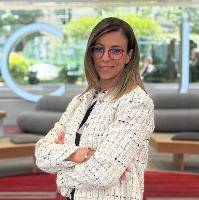 Una mujer de pie con una chaqueta blanca y gafas, sonriendo y con los brazos cruzados en un entorno moderno.