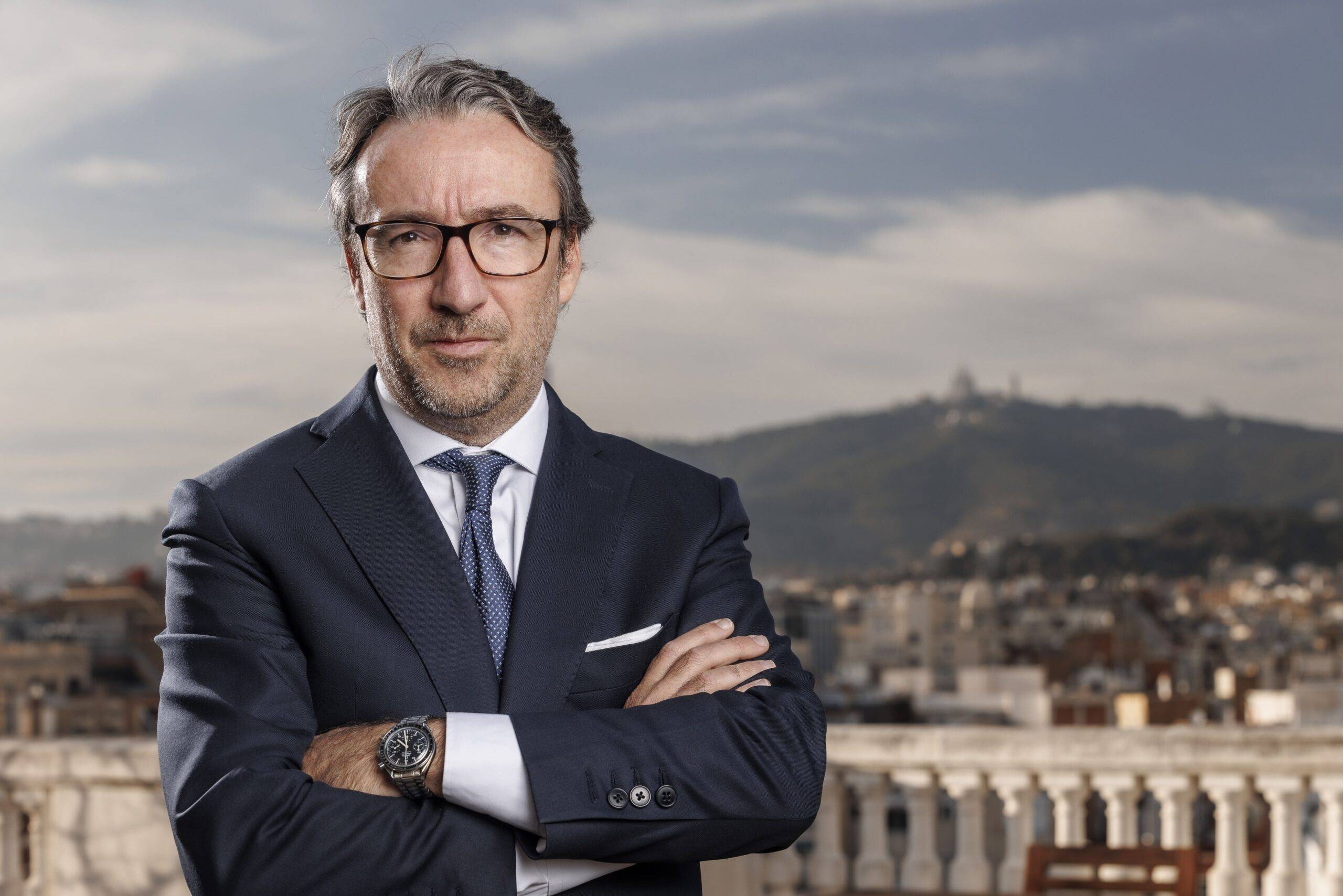 Hombre de negocios con traje y corbata posando con los brazos cruzados frente a un paisaje urbano.