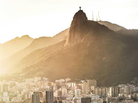Vista aérea de una ciudad con edificios densamente agrupados al pie de una montaña con una estatua en la cima durante el atardecer.