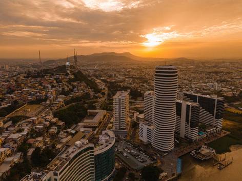 Vista aérea de una ciudad moderna al atardecer, destacando edificios altos y curvos con un fondo de montañas.