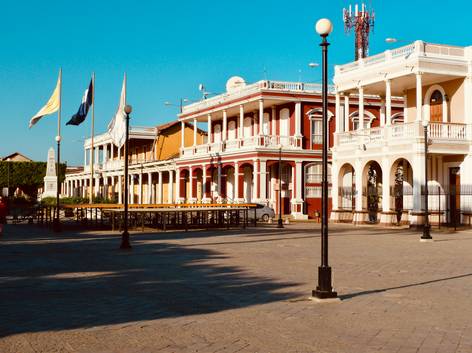 Vista de una plaza pública con edificios de estilo colonial y banderas ondeando.