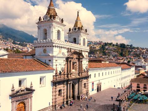 Vista aérea de una iglesia colonial blanca con dos campanarios, ubicada en una plaza con varias personas y casas con tejas rojas en el fondo bajo un cielo parcialmente nublado.