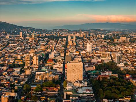 Vista aérea de una ciudad moderna durante el atardecer, mostrando edificios altos y calles transitadas con montañas en el fondo.