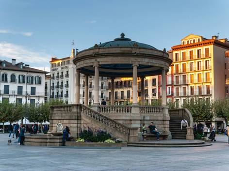 Vista de un quiosco de música en una plaza pública con edificios coloridos alrededor y personas disfrutando del ambiente.