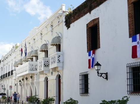 Fachada de edificios coloniales con banderas colgando y personas caminando en una calle soleada.