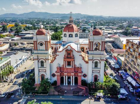 Vista aérea de una catedral de color blanco y rosa con dos cúpulas, ubicada en una zona urbana con edificios circundantes.