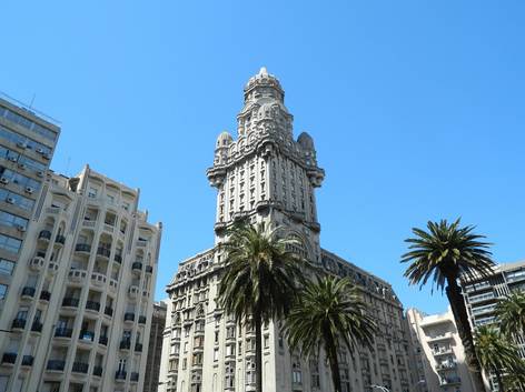 Vista de un imponente edificio en estilo arquitectónico ecléctico con detalles ornamentales, rodeado de palmeras y bajo un cielo despejado.