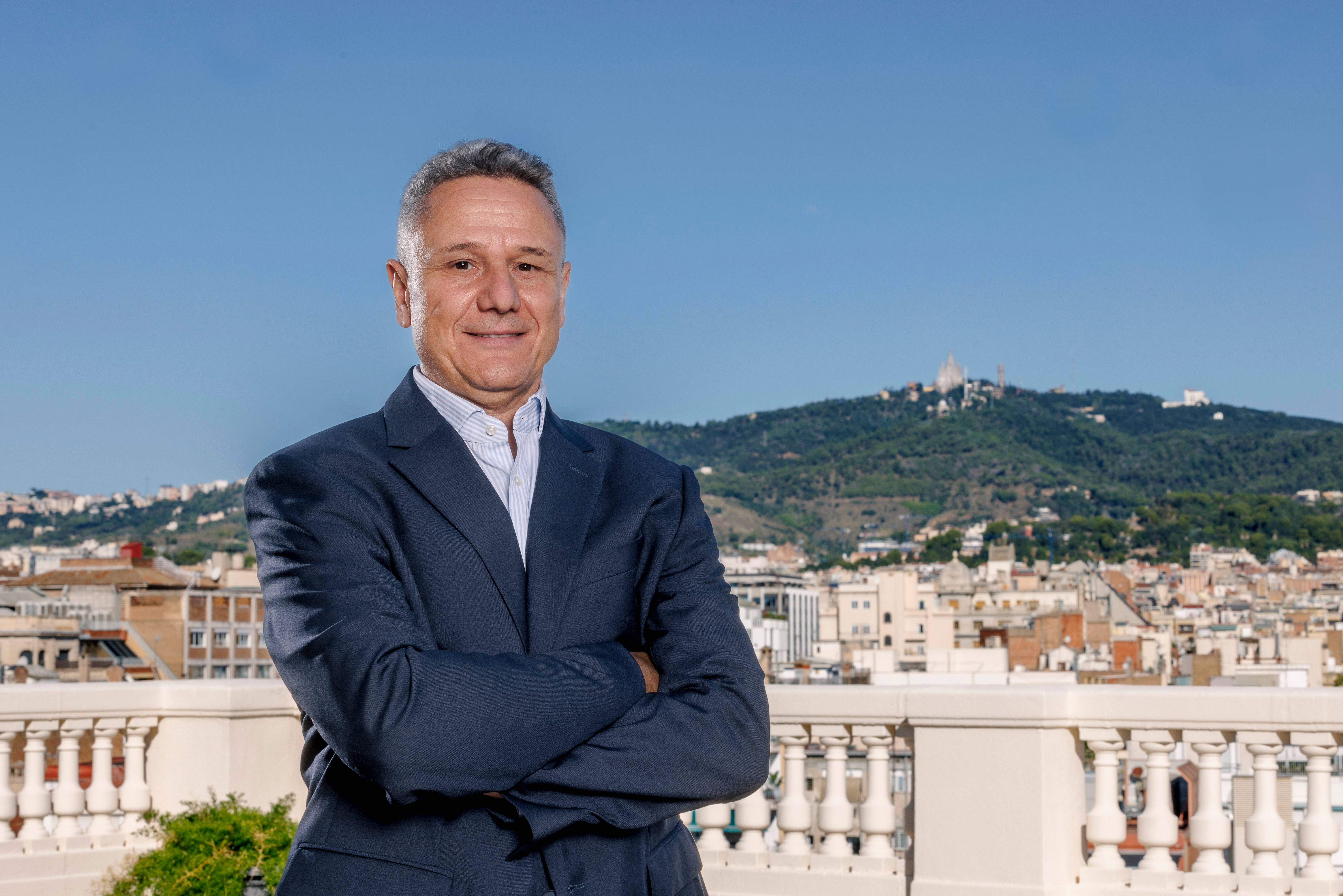 Un hombre de negocios posa con los brazos cruzados frente a una vista de la ciudad con montañas al fondo.
