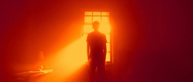 Un hombre de pie delante de una ventana iluminada por un intenso rayo de luz naranja.