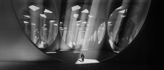 Una figura solitaria camina a través de un vasto espacio arquitectónico iluminado por rayos de luz que entran a través de ventanas rectangulares.