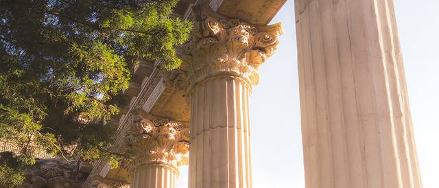 La imagen muestra columnas clásicas de estilo griego, con decoración elaborada y un fondo natural.