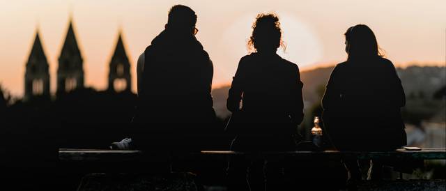 Tres personas sentadas en un banco contemplando un atardecer con torres al fondo.