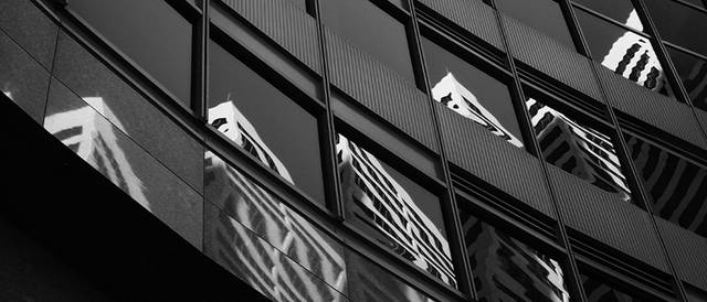 La imagen muestra reflejos de edificios en las ventanas de una estructura moderna, capturada en blanco y negro.