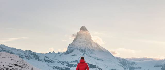 Una persona de espaldas, vistiendo una chaqueta roja, observa un majestuoso pico nevado al atardecer.