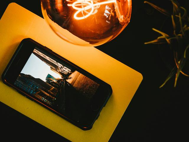 Un teléfono móvil está apoyado sobre una superficie amarilla con una lámpara de luz cálida y un fondo oscuro.
