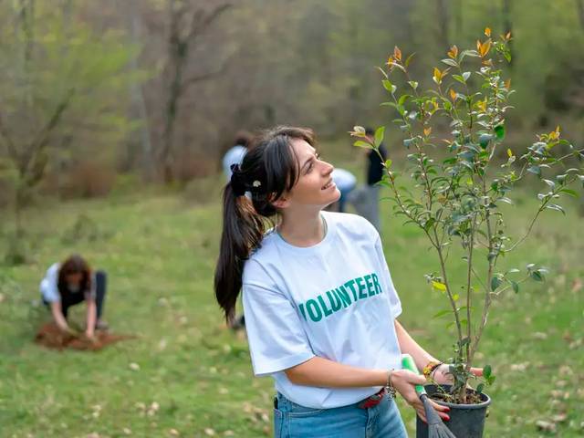 Una voluntaria sonriente sostiene una planta en un entorno natural mientras otros voluntarios trabajan en el fondo.