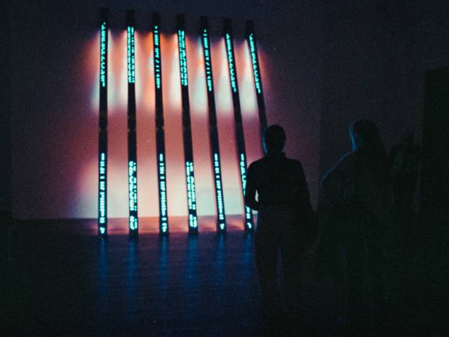 La imagen presenta una instalación artística con luces coloridas en una pared, mientras dos personas observan desde el primer plano.