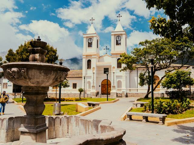 La imagen muestra una plaza con una fuente ornamental y una iglesia al fondo, rodeada de árboles y un cielo despejado.