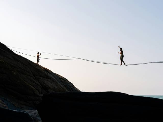 Dos personas caminan sobre una cuerda floja entre rocas junto al mar.