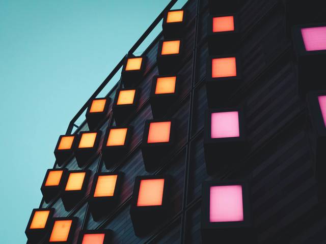 La imagen muestra una pared con paneles iluminados en colores naranja, rosa y blanco contra un fondo azul claro.