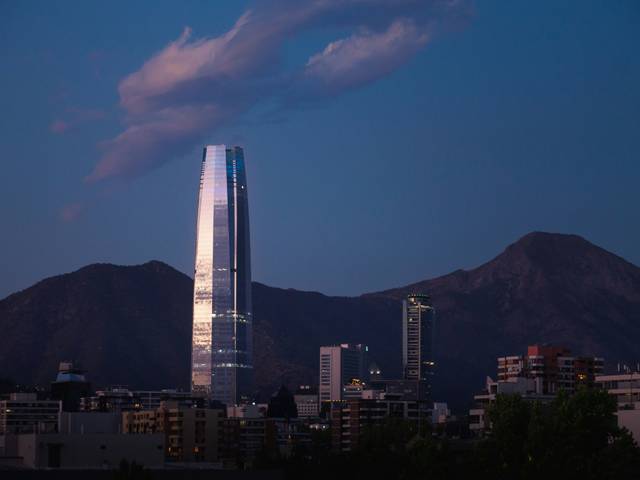 La imagen muestra un rascacielos moderno iluminado al atardecer en una ciudad rodeada de montañas.