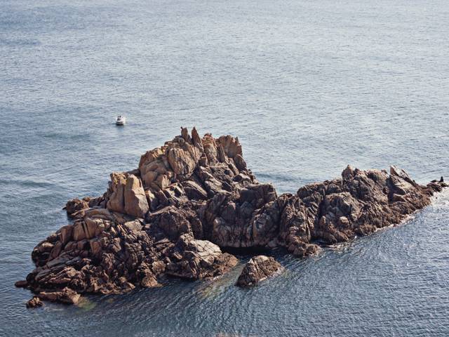 Una isla rocosa en medio del mar con un pequeño barco al fondo.