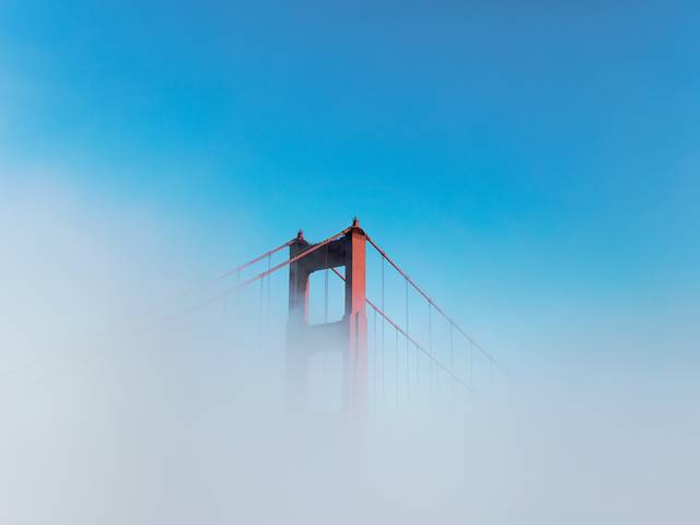 Un puente rojo parcialmente visible a través de la niebla bajo un cielo azul.