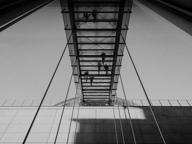 La imagen muestra un puente colgante de cristal con la sombra proyectada sobre una estructura moderna.