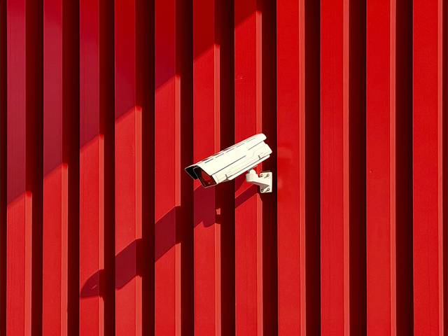La imagen muestra una cámara de seguridad montada sobre una pared roja con líneas verticales.