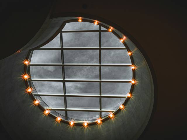 Una vista desde abajo de un tragaluz redondo con unas luces alrededor y nubes visibles en el cielo.