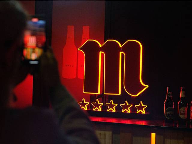 La imagen muestra un letrero iluminado con la letra 'm' y varias estrellas, con botellas de cerveza en el fondo.