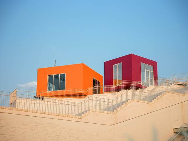 Dos casas coloridas, una naranja y otra morada, se encuentran en una colina con un cielo azul claro de fondo.