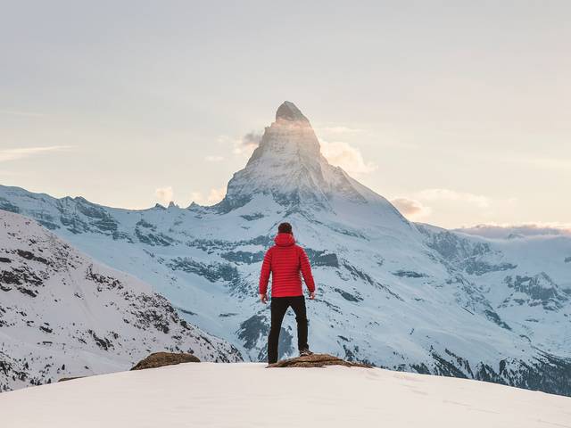 Una persona de espaldas, vistiendo una chaqueta roja, observa un majestuoso pico nevado al atardecer.